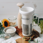 VeganMilker® SOUL с дървено бутало стои на маса, заобиколена от купички с ориз, семена и слънчоглед.