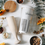 VeganMilker® SOUL с дървена шпатула и разнообразни ядки върху бяла повърхност, акцентирани със слънчогледи и листа.